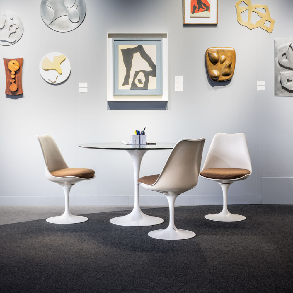 Mehrere Kunstbilder und Objekte hängen an einer grauen Wand, davor steht ein weisser, runder Pantontisch und 3 weisse Pantonstühle.