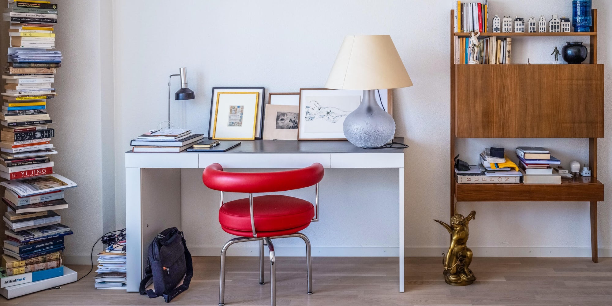 Ein Schreibtisch mit einem roten, auffallenden Designerstuhl, links und rechts Regale mit Bücher und Deko. Auf dem Tisch stehen bei Tischlampen. 